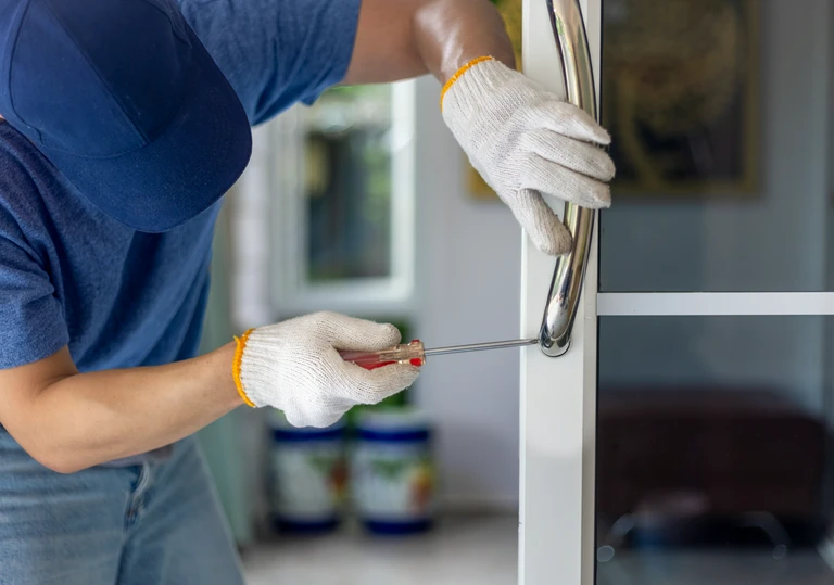 Pracownik przykręca śrubokrętem długi, metalowy uchwyt (pochwyt) do&nbsp;białych drzwi